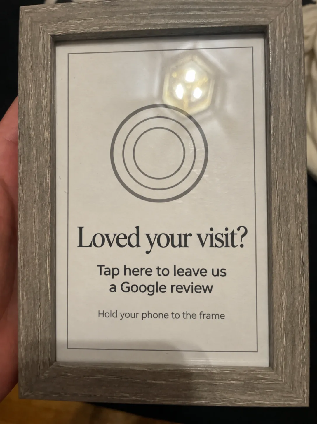 The TapIt Frame held in a hand: grey weathered-wood frame, cream insert with a tap mark and the words Loved your visit? Tap here to leave us a Google review